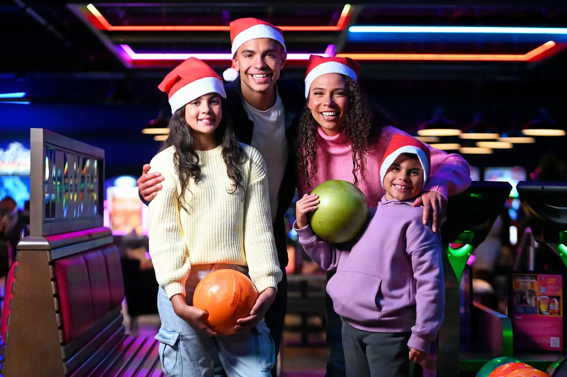 A group of four people wearing Santa hats, holding bowling balls, poses joyfully in a vibrant bowling alley with colorful lights.