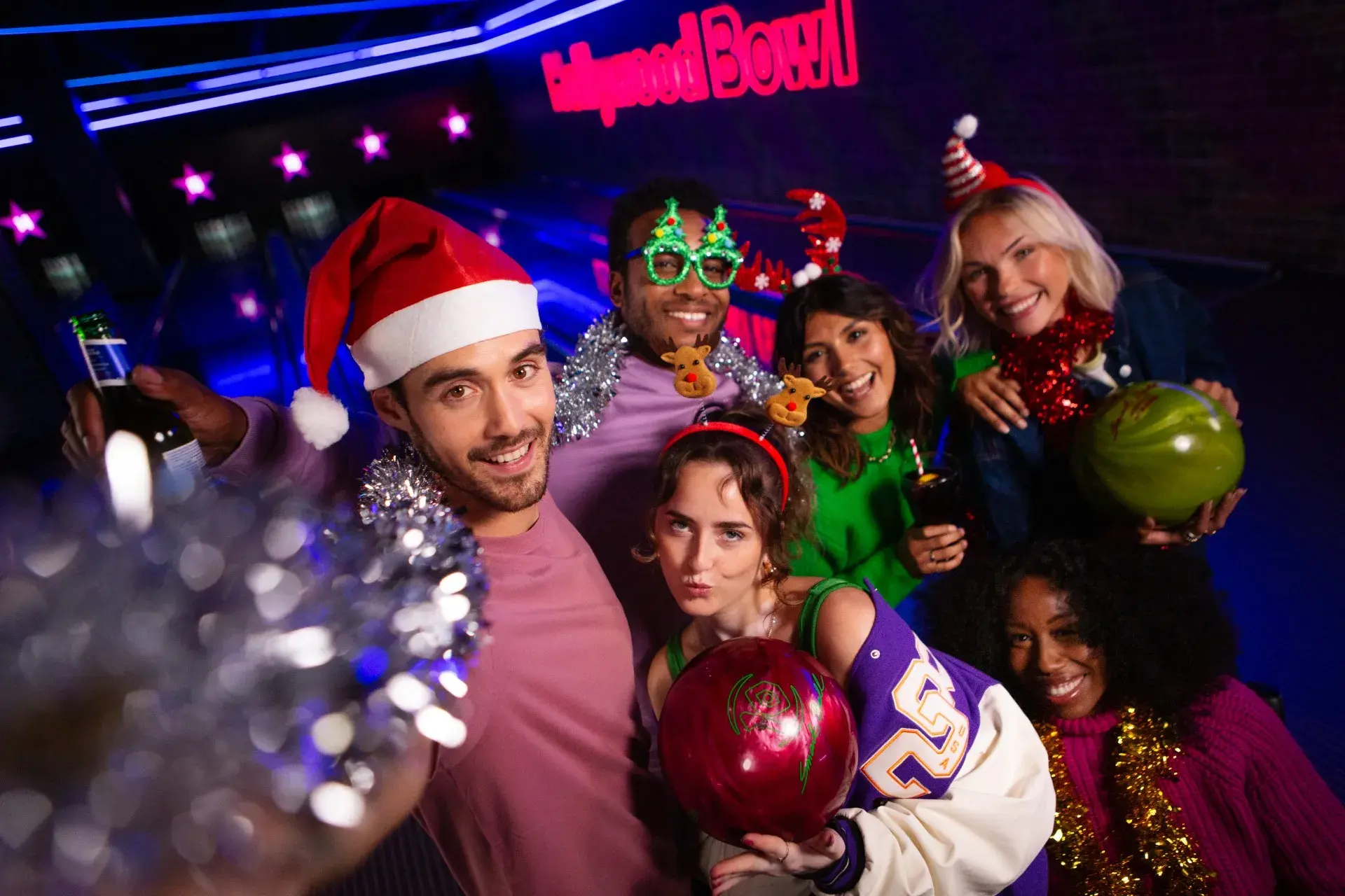 A joyful group of four people wearing Santa hats hold bowling balls and pose cheerfully in a brightly lit bowling alley, conveying holiday fun.