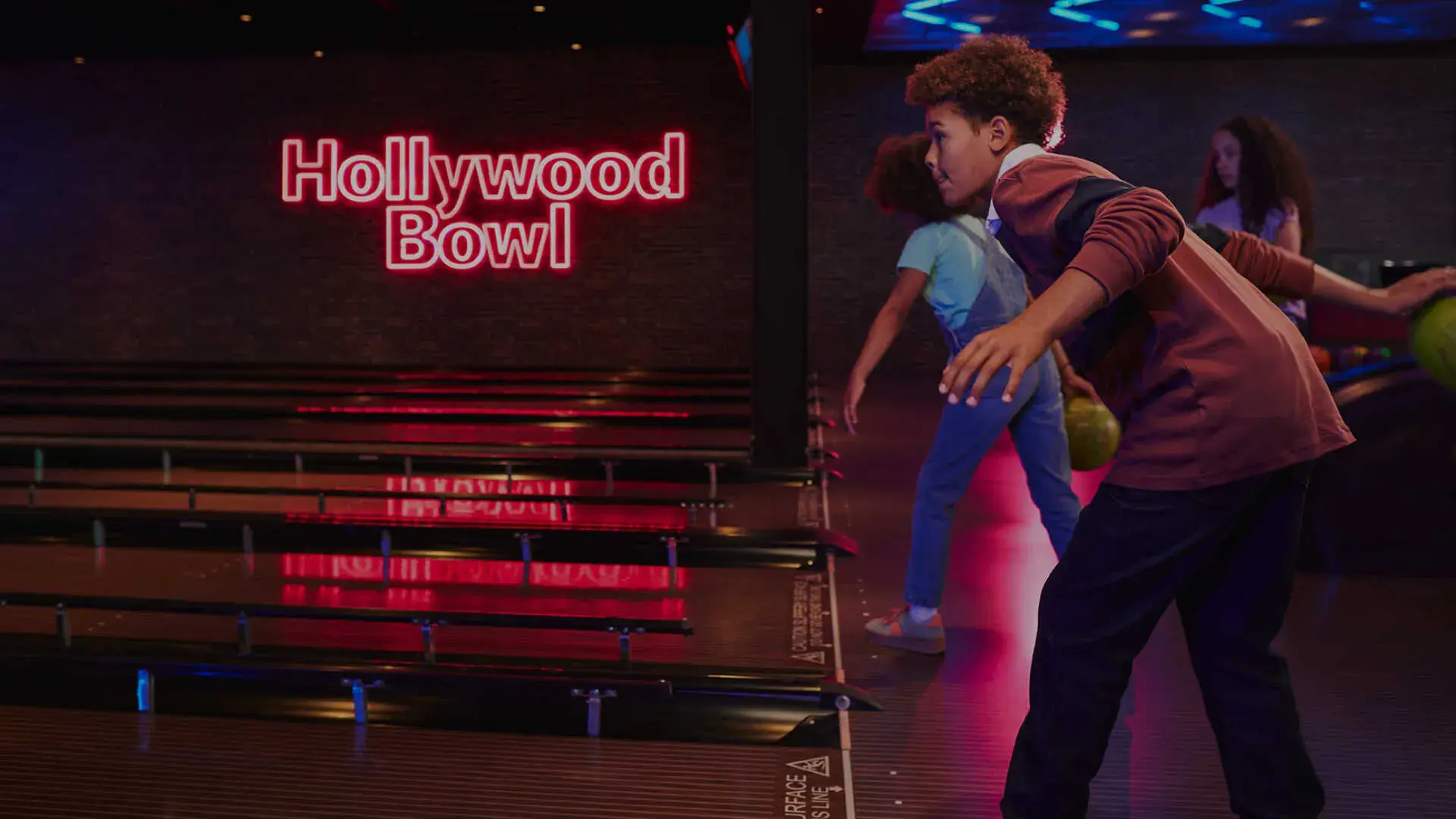 A young boy in a casual outfit bowls at a vibrant alley under a neon "Hollywood Bowl" sign. Two friends bowl nearby, creating a fun, energetic atmosphere.