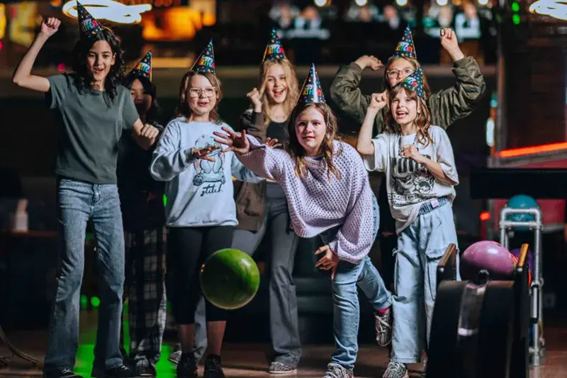 A group of kids at a bowling alley, all wearing colourful party hats, watch excitedly as one of them rolls a green bowling ball.