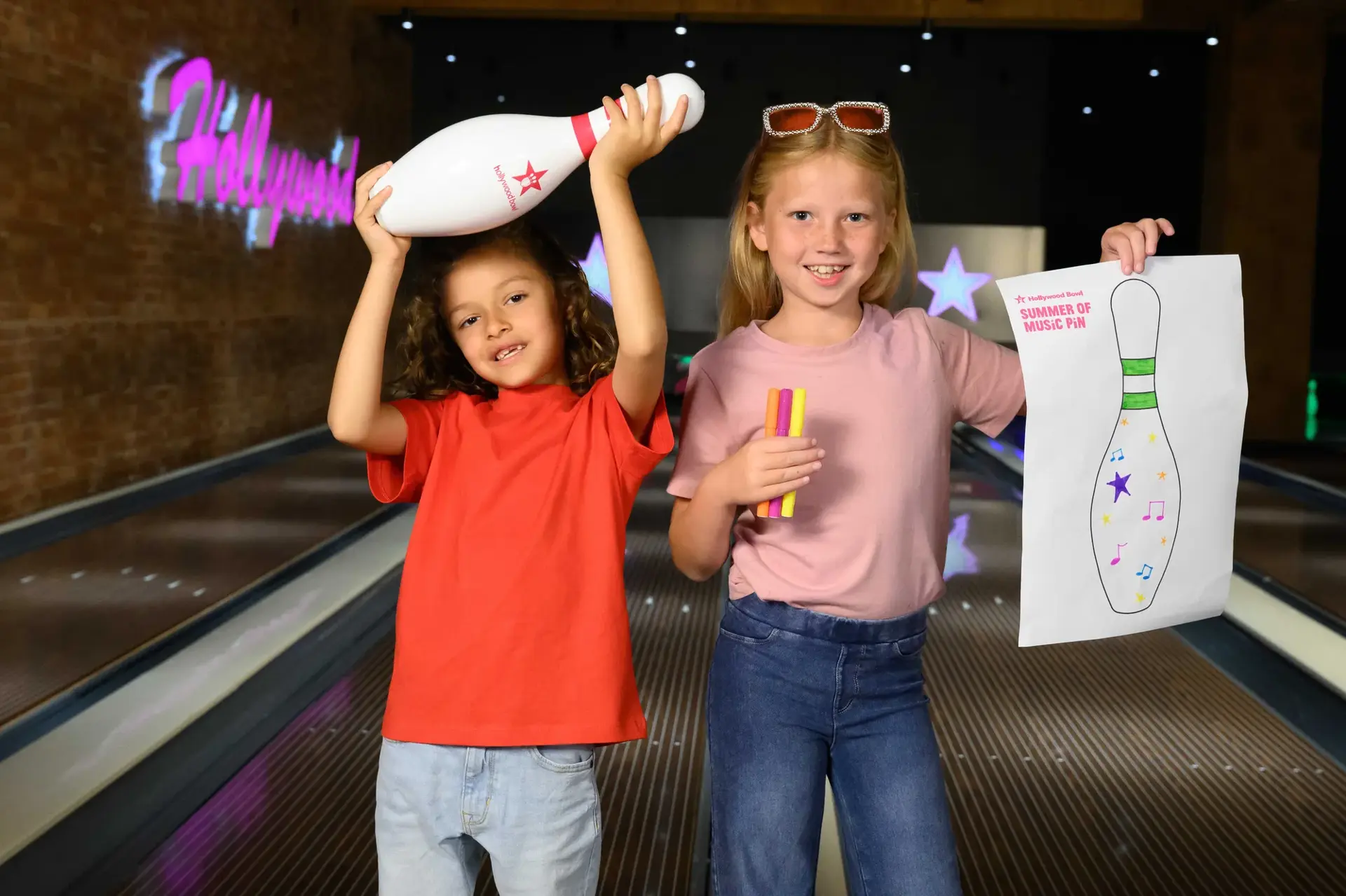 Two smiling children display bowling pins and a colourful sign, showcasing their excitement in a fun bowling environment.