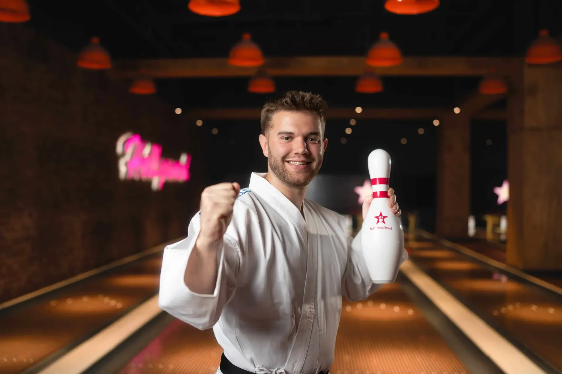 A man wearing Karate robes smiles while holding a bowling pin, ready to take his turn at the bowling alley.