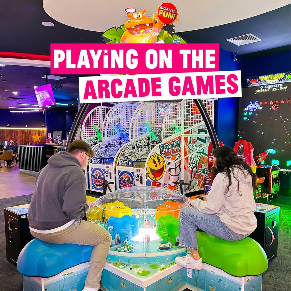Two people sit facing each other, playing an arcade game in a brightly lit gaming center with colourful machines around them.