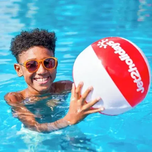 A young boy smiles in a pool while holding a vibrant red and white beach ball above the water.