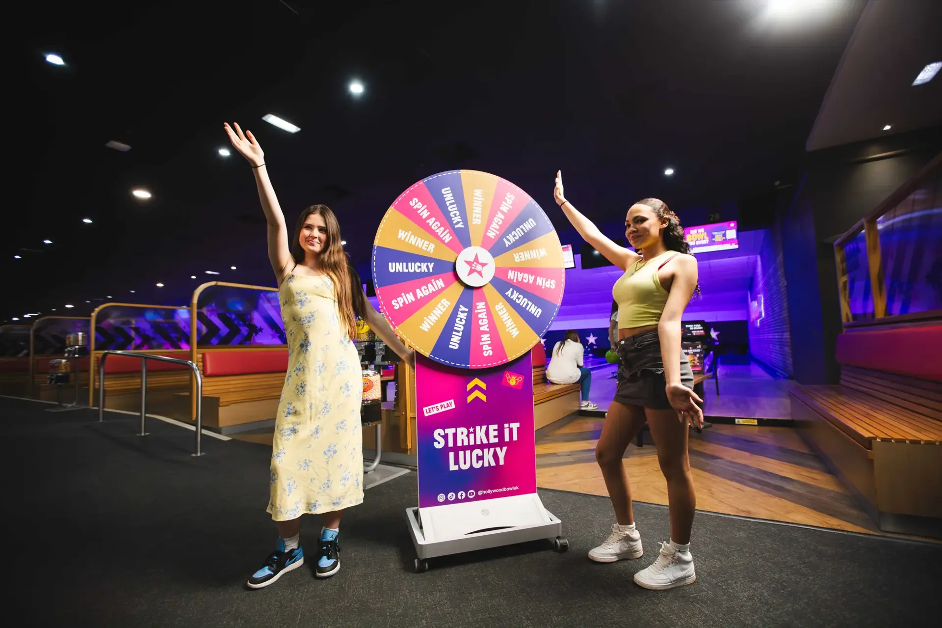 Two women stand beside a spinning wheel, engaged in conversation, with the wheel prominently displayed in the foreground.