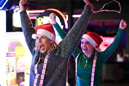 Two friends in Santa hats enjoying a game of pinball, sharing laughter and holiday cheer in a festive atmosphere.