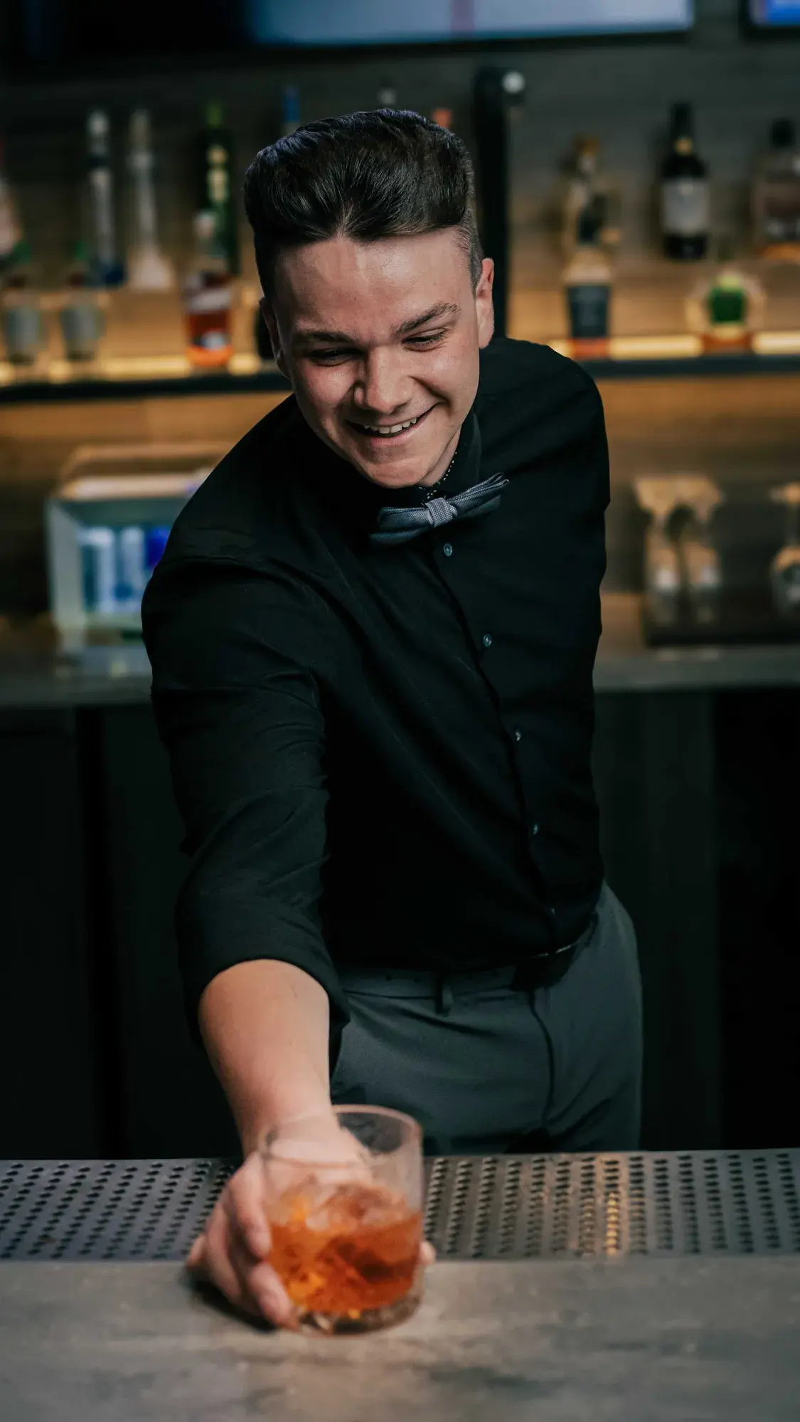 A man in a black shirt raises a glass of whiskey, exuding a relaxed and friendly vibe in a social setting.