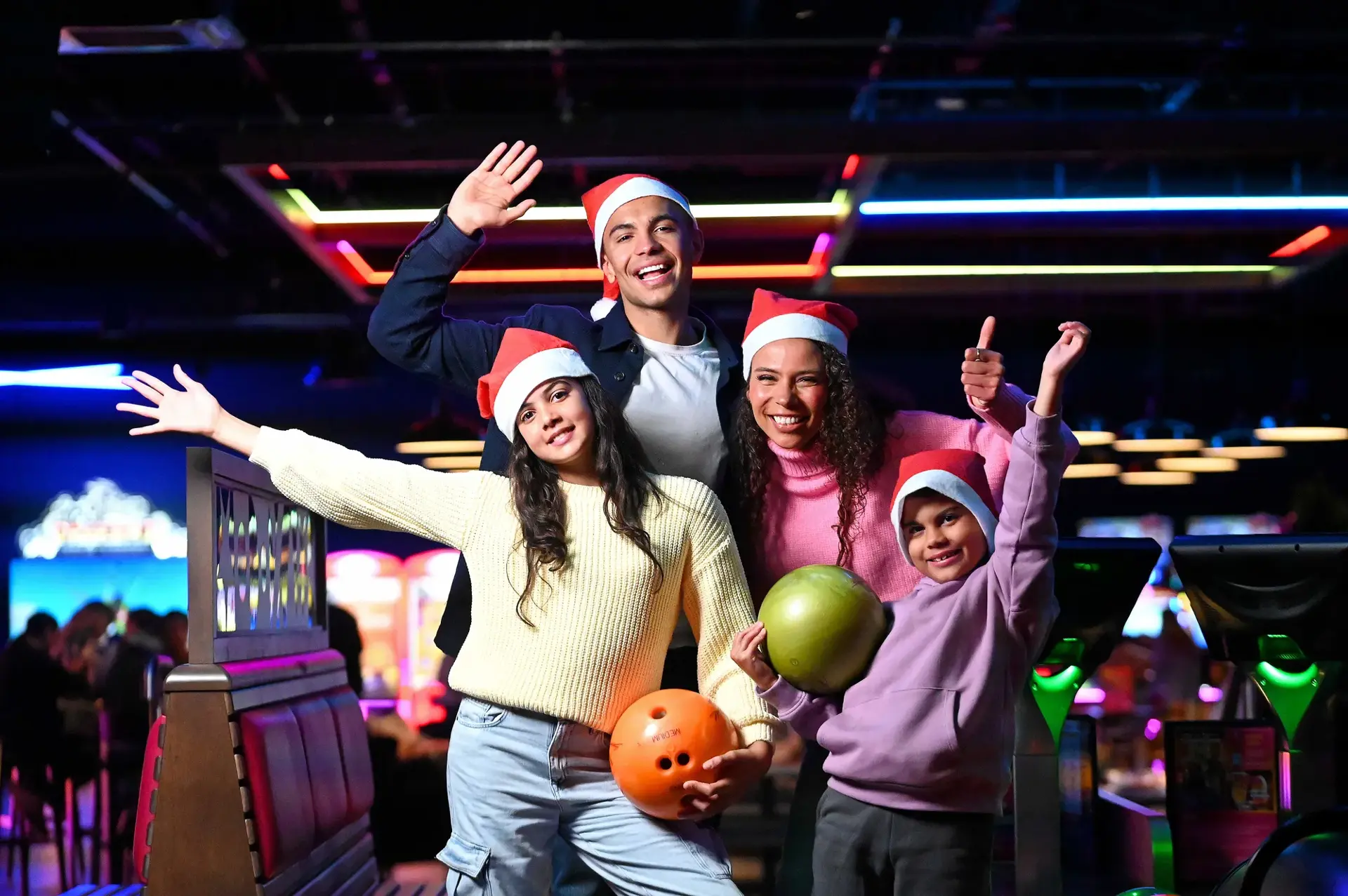A joyful group of four people wearing Santa hats hold bowling balls and pose cheerfully in a brightly lit bowling alley, conveying holiday fun.